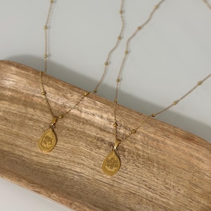 May include: Two gold necklaces with teardrop pendants, each with an embossed design. The necklaces are displayed on a wooden tray. The chains have small, round beads. The pendants feature a detailed, embossed pattern.