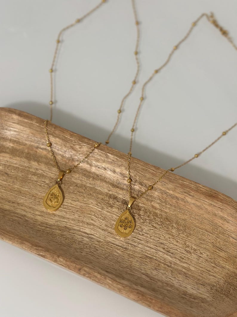 May include: Two gold necklaces with teardrop pendants, each with an embossed design. The necklaces are displayed on a wooden tray. The chains have small, round beads. The pendants feature a detailed, embossed pattern.