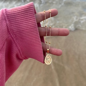 May include: A gold-colored necklace with a teardrop pendant featuring an engraved design. The necklace has a delicate chain with small, evenly spaced beads. The background shows a pink garment and the ocean.