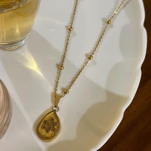 May include: Gold-coloured necklace with a teardrop pendant. The pendant has an intricate design within a textured frame. The chain includes small, evenly spaced beads. The necklace is displayed on a white, scalloped-edge plate.