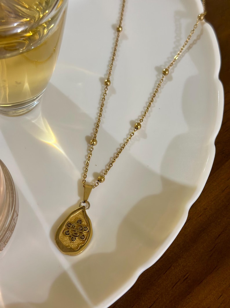 May include: Gold-coloured necklace with a teardrop pendant. The pendant has an intricate design within a textured frame. The chain includes small, evenly spaced beads. The necklace is displayed on a white, scalloped-edge plate.