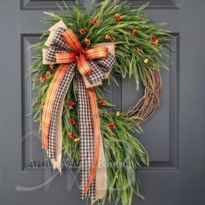 May include: A wreath featuring green foliage and small orange flowers on a brown twig base. A large decorative bow is attached, made of layered ribbons in tan burlap, black and white buffalo check, and an orange, red, and yellow plaid pattern.