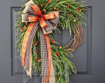 May include: A wreath featuring green fern-like foliage and small orange flowers on a brown twig base. A large decorative bow made of burlap, black and white gingham, and orange plaid ribbon hangs from the top left, with long trailing ribbon tails.
