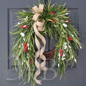 May include: A wreath featuring green fern-like foliage, small red and white flowers, and a central tan burlap ribbon bow with long trailing tails. The wreath is constructed on a brown twig base and includes the text Artful Wreath Boutique.