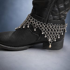 May include: A black boot featuring a quilted upper section and a side zipper, adorned with a silver-toned chainmail ankle accessory. The accessory consists of interlocking metal rings with small dangling beads along the bottom edge.