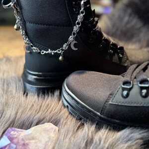 May include: Black combat boots with silver-tone chain accents, including crescent moon and bell charms. The boots have a textured fabric upper and a thick, black sole. The boots are displayed on a faux fur surface.