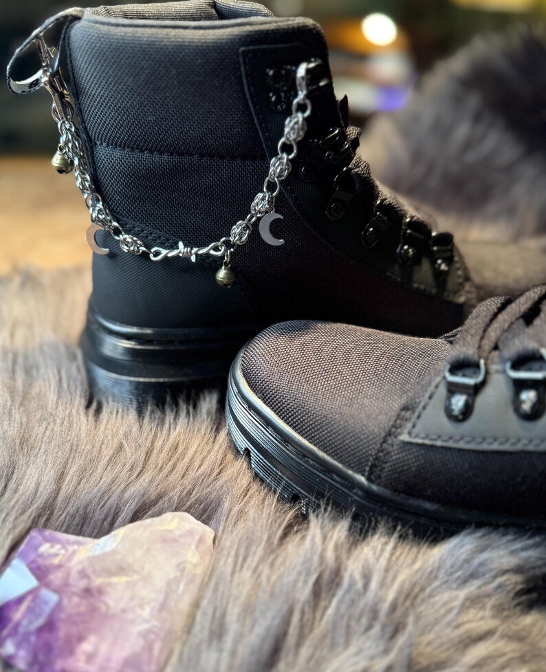 May include: Black combat boots with silver-tone chain accents, including crescent moon and bell charms. The boots have a textured fabric upper and a thick, black sole. The boots are displayed on a faux fur surface.