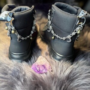 May include: Pair of black ankle boots with silver chain and charm accents, including crescent moons and bells. The boots have a zipper closure and are set on a gray, furry surface. A purple and white crystal is placed between the boots.