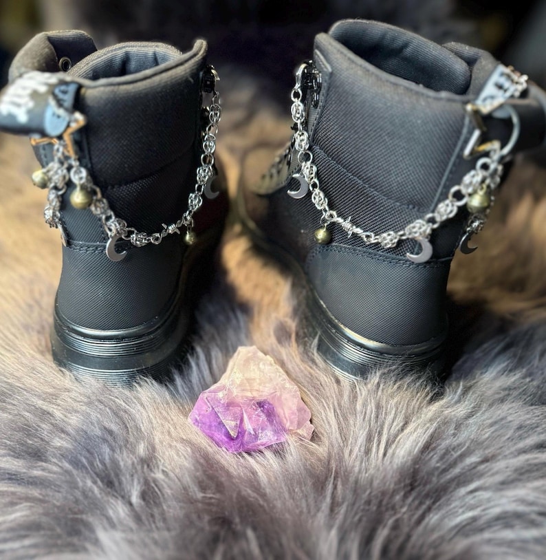 May include: Pair of black ankle boots with silver chain and charm accents, including crescent moons and bells. The boots have a zipper closure and are set on a gray, furry surface. A purple and white crystal is placed between the boots.