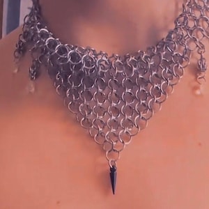 May include: A silver chainmail necklace with a triangular design. The necklace has a series of small, interlocking metal rings. The necklace has a black spike pendant and small dangling beads. The jewelry is designed to be worn around the neck.