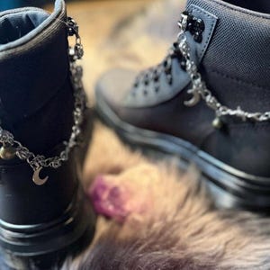 May include: A pair of black ankle boots with silver-colored chain accents. The chains feature small charms, including crescent moons and beads. The boots have a matte finish and are displayed on a textured surface.