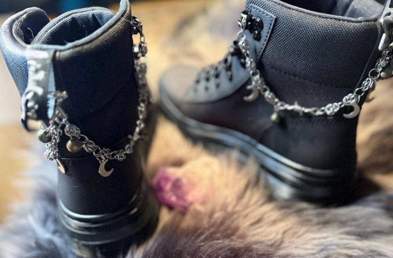 May include: A pair of black ankle boots with silver-colored chain accents. The chains feature small charms, including crescent moons and beads. The boots have a matte finish and are displayed on a textured surface.