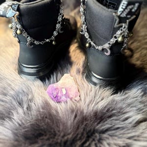 May include: Pair of black ankle boots with silver-tone chain and charm accents, including moons and stars. The boots are set on a dark surface with a purple crystal in the center.