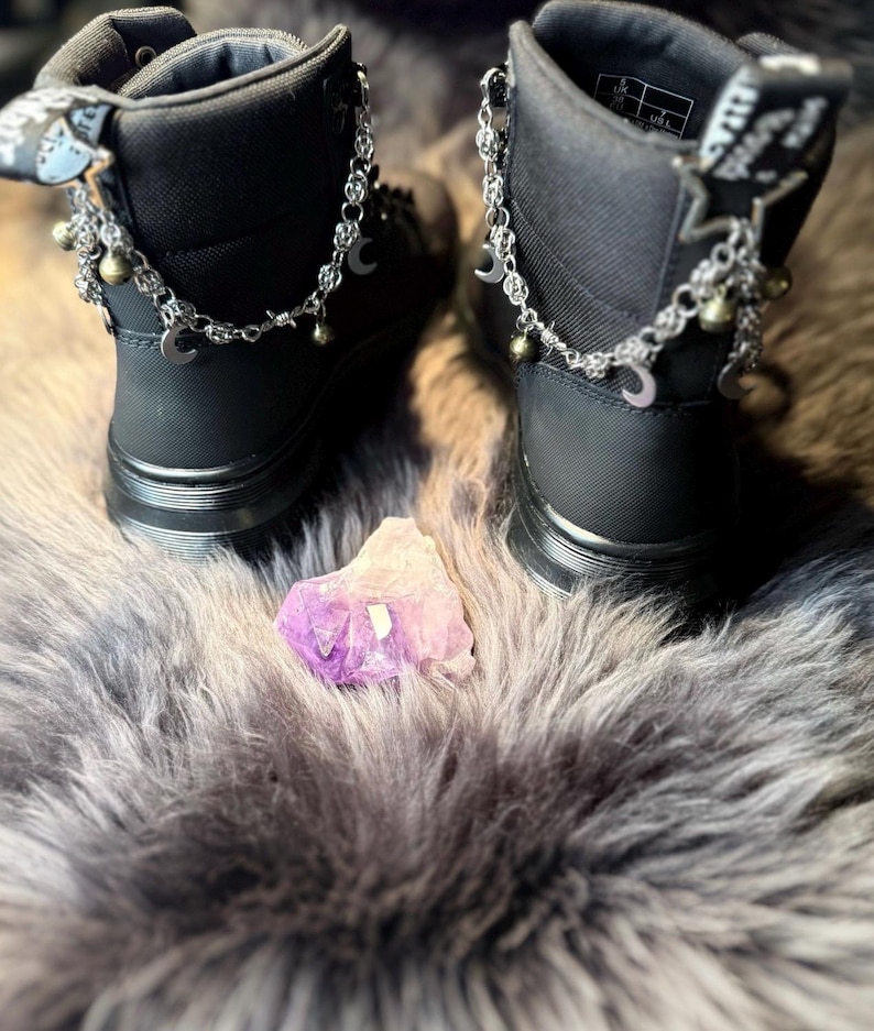 May include: Pair of black ankle boots with silver-tone chain and charm accents, including moons and stars. The boots are set on a dark surface with a purple crystal in the center.