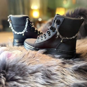 May include: Black combat boots with silver chain accents and decorative charms. The boots have a lace-up design and a thick sole. The boots are displayed on a faux fur surface.