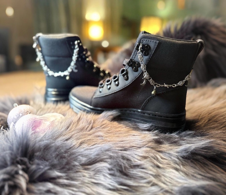 May include: Black combat boots with silver chain accents and decorative charms. The boots have a lace-up design and a thick sole. The boots are displayed on a faux fur surface.