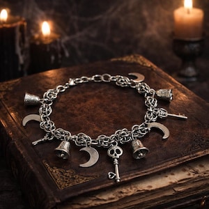 May include: Silver charm bracelet featuring crescent moons, bells, and a skull key. The bracelet is displayed on a dark, textured surface with lit candles in the background, creating a mystical aesthetic.