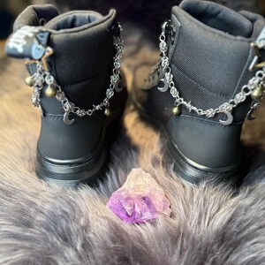 May include: A pair of black ankle boots with silver chain accents, crescent moon charms, and small bells. A purple and white crystal sits between the boots. The boots have zippers and a textured fabric.