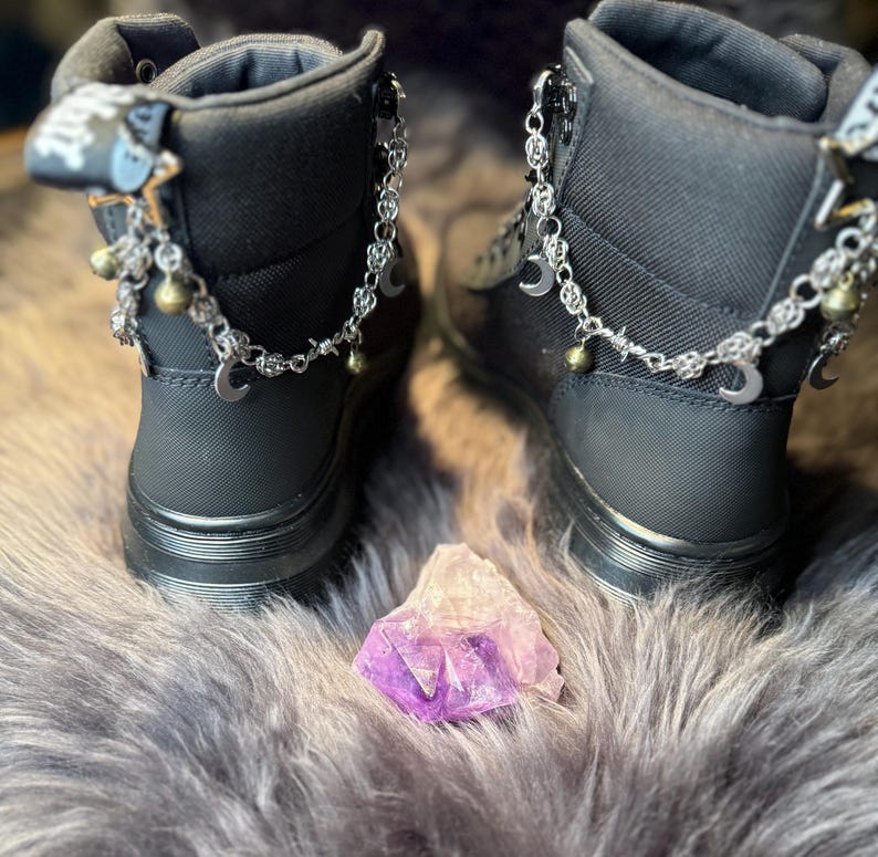 May include: A pair of black ankle boots with silver chain accents, crescent moon charms, and small bells. A purple and white crystal sits between the boots. The boots have zippers and a textured fabric.