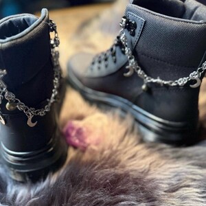 May include: A pair of black ankle boots with silver-colored chain accents. The chains feature small charms, including crescent moons and bells. The boots are set on a faux fur surface.