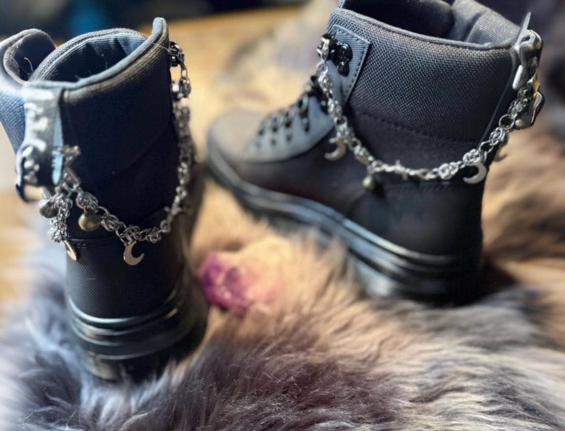 May include: A pair of black ankle boots with silver-colored chain accents. The chains feature small charms, including crescent moons and bells. The boots are set on a faux fur surface.