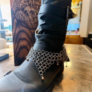 May include: A black leather boot with a silver chainmail anklet. The anklet has a geometric design with small gold-colored beads hanging from the bottom. A wooden box with a skull carving is in the background.