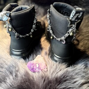 May include: A pair of black ankle boots with silver chain and charm accents. The chains feature crescent moons and small bells. A purple and white crystal sits between the boots on a gray, furry surface.