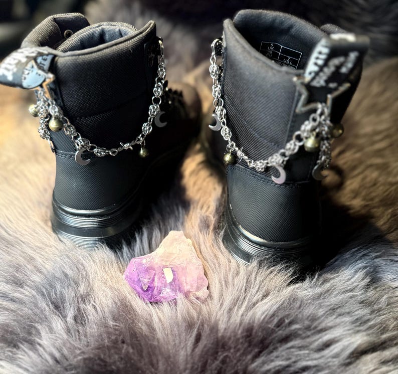 May include: A pair of black ankle boots with silver chain and charm accents. The chains feature crescent moons and small bells. A purple and white crystal sits between the boots on a gray, furry surface.