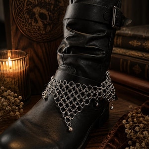 May include: A black leather slouch boot featuring a silver-toned chainmail accessory wrapped around the ankle. The chainmail is constructed from interlocking metal rings in a triangular pattern, with small spherical charms hanging from the bottom edge.