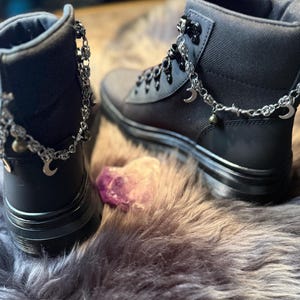 May include: Black ankle boots with silver-tone chain accents, including crescent moons and beads. The boots have a matte finish and are laced up. The image shows the boots on a faux fur surface, with a purple crystal in the foreground.
