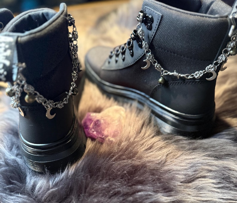 May include: Black ankle boots with silver-tone chain accents, including crescent moons and beads. The boots have a matte finish and are laced up. The image shows the boots on a faux fur surface, with a purple crystal in the foreground.