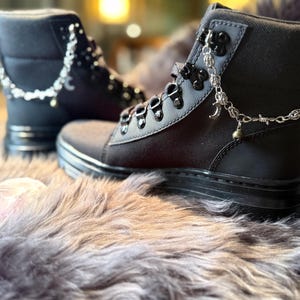 May include: Black combat boots with silver chain accents. The boots have a sturdy sole, a lace-up design, and a decorative chain with charms. The boots are made of a durable material and are suitable for various occasions.