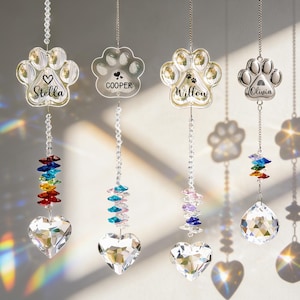 May include: Four hanging crystal suncatchers with paw print designs. Each suncatcher has a different name: Stella, Cooper, Willow, and Olivia. The suncatchers have a heart-shaped crystal at the bottom and a rainbow-colored crystal accent.