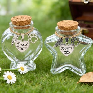 Könnte beinhalten: Zwei durchsichtige Glas-Andenken-Gläser mit Korken. Eines ist herzförmig, das andere sternförmig. Jedes hat einen Anhänger mit einem Flügel, einem Pfotenabdruck und einem Namen: "Basil" und "Cooper".