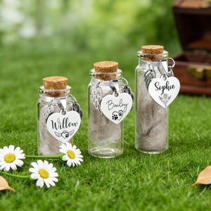 May include: Three small glass bottles with cork stoppers, each containing a fluffy substance. Each bottle has a silver heart-shaped charm with a name engraved on it: Willow, Bailey, and Sophie. The bottles are on green grass.