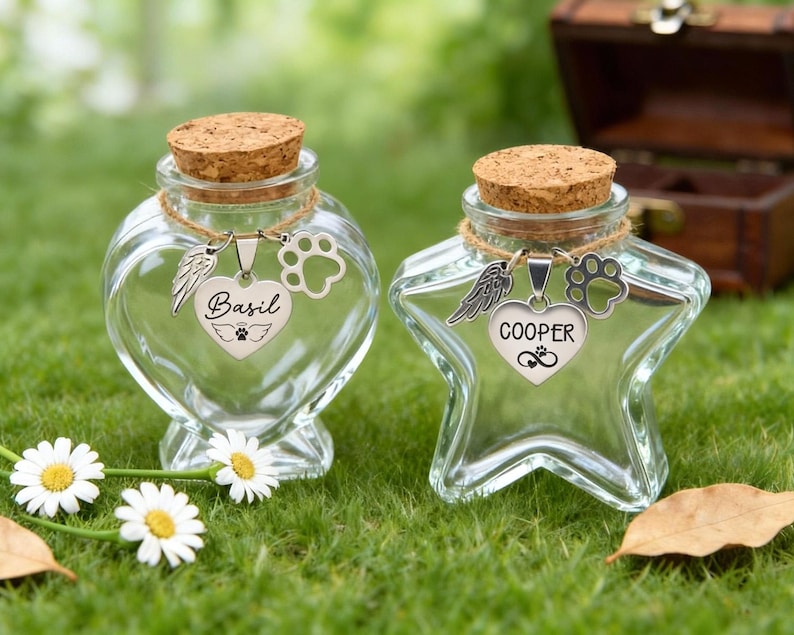 May include: Two clear glass keepsake jars with cork stoppers. One is heart-shaped, the other star-shaped. Each has a charm with a wing, paw print, and a name: "Basil" and "Cooper". The jars are on green grass with white daisies.