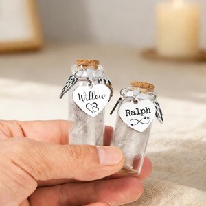 May include: Two miniature glass bottles with cork stoppers, each filled with a fluffy material. One bottle features a heart-shaped tag reading "Willow" with two hearts, the other reads "Ralph" with a paw print and infinity symbol. Both have silver wings.