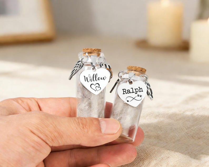 May include: Two miniature glass bottles with cork stoppers, each filled with a fluffy material. One bottle features a heart-shaped tag reading "Willow" with two hearts, the other reads "Ralph" with a paw print and infinity symbol. Both have silver wings.
