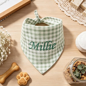May include: A sage green and white gingham patterned pet bandana featuring the name Millie embroidered in dark green cursive script. The triangular fabric accessory has a knotted top section designed to be tied around a pet's neck.