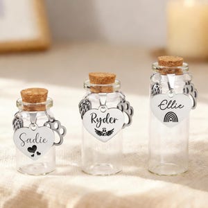 May include: Three small glass bottles with cork stoppers and heart-shaped tags. The tags are engraved with names and symbols: "Sadie" with hearts, "Ryder" with wings, and "Ellie" with a rainbow. The bottles are clear, and the tags are silver.