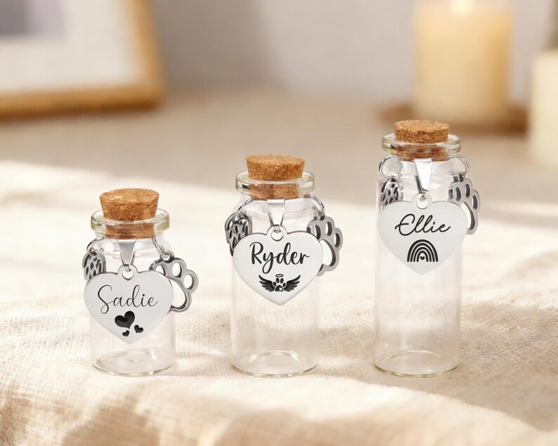 May include: Three small glass bottles with cork stoppers and heart-shaped tags. The tags are engraved with names and symbols: "Sadie" with hearts, "Ryder" with wings, and "Ellie" with a rainbow. The bottles are clear, and the tags are silver.