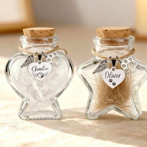 May include: Two clear glass keepsake jars with cork stoppers. One is heart-shaped, the other star-shaped. Each jar has a silver charm with a name and paw print. The heart jar says "Charlie", the star jar says "Oliver".