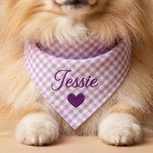 May include: A triangular pet bandana featuring a light purple and white gingham check pattern. The fabric is embroidered with the name Jessie in a dark purple cursive script, positioned directly above a solid dark purple heart icon.