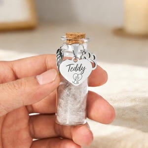 May include: A small glass vial with a cork stopper, filled with a light-coloured substance. A silver heart-shaped charm with the name "Teddy" and a paw print hangs from the top. The vial is designed to hold a pet's ashes.