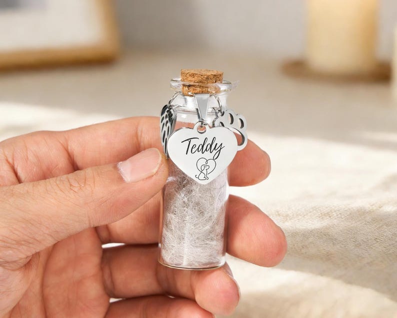 May include: A small glass vial with a cork stopper, filled with a light-coloured substance. A silver heart-shaped charm with the name "Teddy" and a paw print hangs from the top. The vial is designed to hold a pet's ashes.