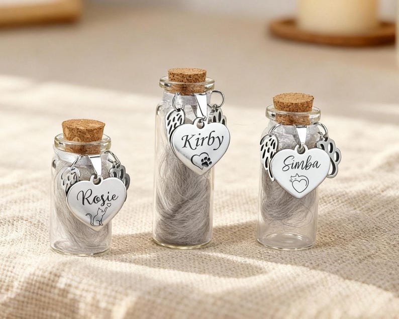 May include: Three small glass bottles with cork stoppers, each containing a pet's ashes. Each bottle has a silver heart-shaped charm with a pet's name and a pet-related design. The bottles are displayed on a neutral-coloured surface.