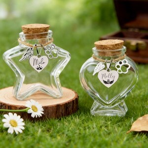 May include: Two clear glass keepsake jars with cork stoppers. One is star-shaped, the other heart-shaped. Each has a charm with a name and paw print. The star jar says "Milo", the heart jar says "Molly". Both sit on a wooden slice.