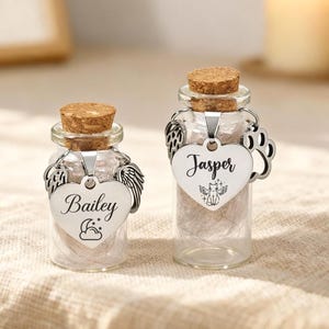 May include: Two small glass bottles with cork stoppers. Each bottle has a silver heart-shaped tag with a name and a decorative charm. One tag says "Bailey" with angel wings, the other says "Jasper" with a paw print.