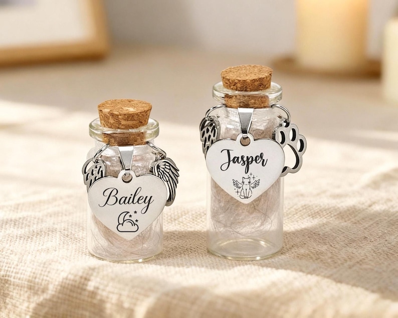 May include: Two small glass bottles with cork stoppers. Each bottle has a silver heart-shaped tag with a name and a decorative charm. One tag says "Bailey" with angel wings, the other says "Jasper" with a paw print.