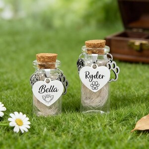 May include: Two small glass bottles with cork stoppers, filled with a light-coloured material. Each bottle has a heart-shaped tag with a name and paw print. One says "Bella" and the other says "Ryder".